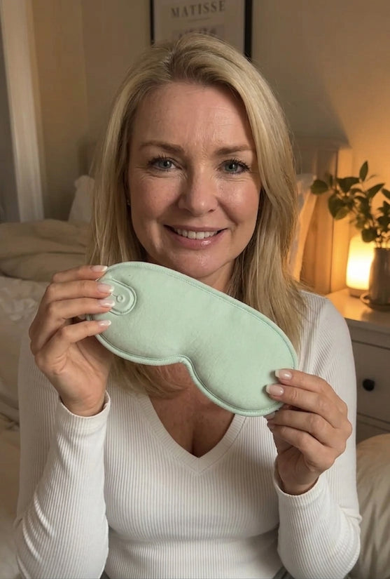 Woman sitting on a bed at night with her eyes closed, holding the green heated eye mask